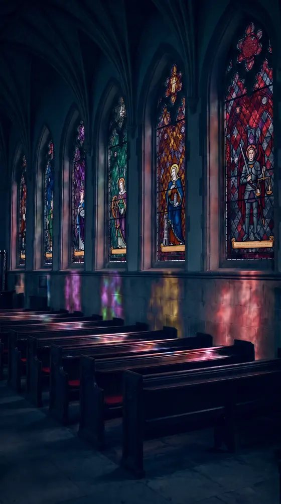 A row of stained-glass saints in a dark gothic chapel, coloured light on the pews